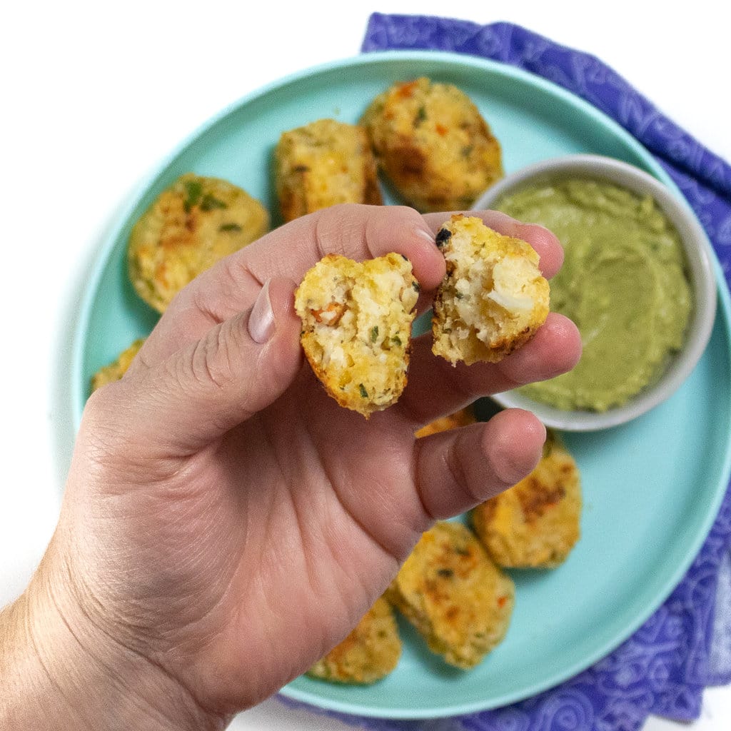 Hand holding up a cut in half cauliflower tots to show the insides against a plate of cauliflower tots in a white background.