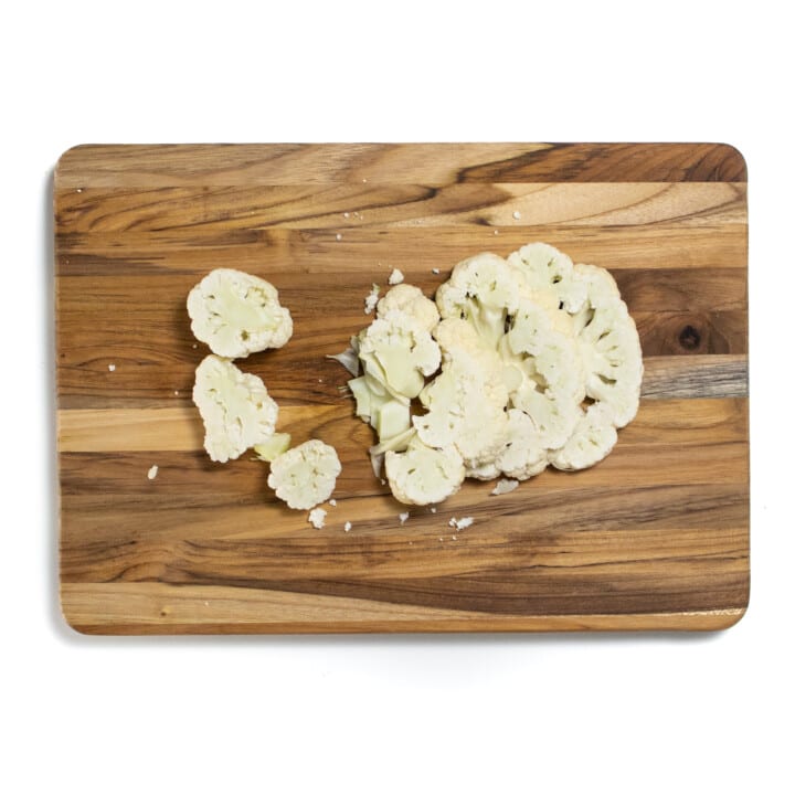 On a wooden cutting board there are steaks of cauliflower laying there.