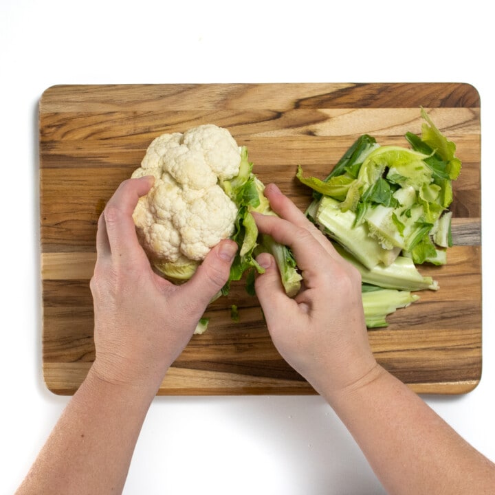 Wooden cutting board with two hands taking the leaves of a head of cauliflower.