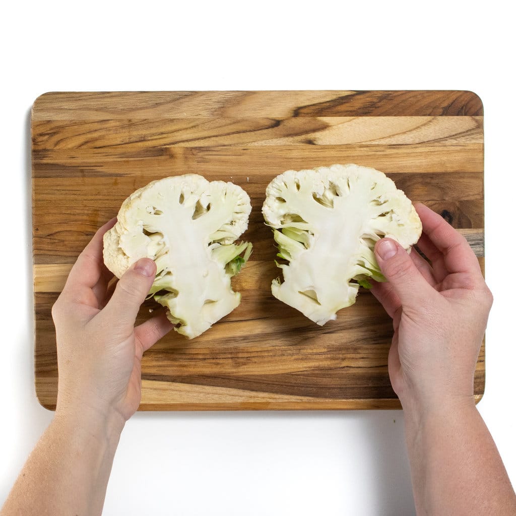 On a wooden cutting board a color flower head is cut in half with two hands holding a cauliflower up.