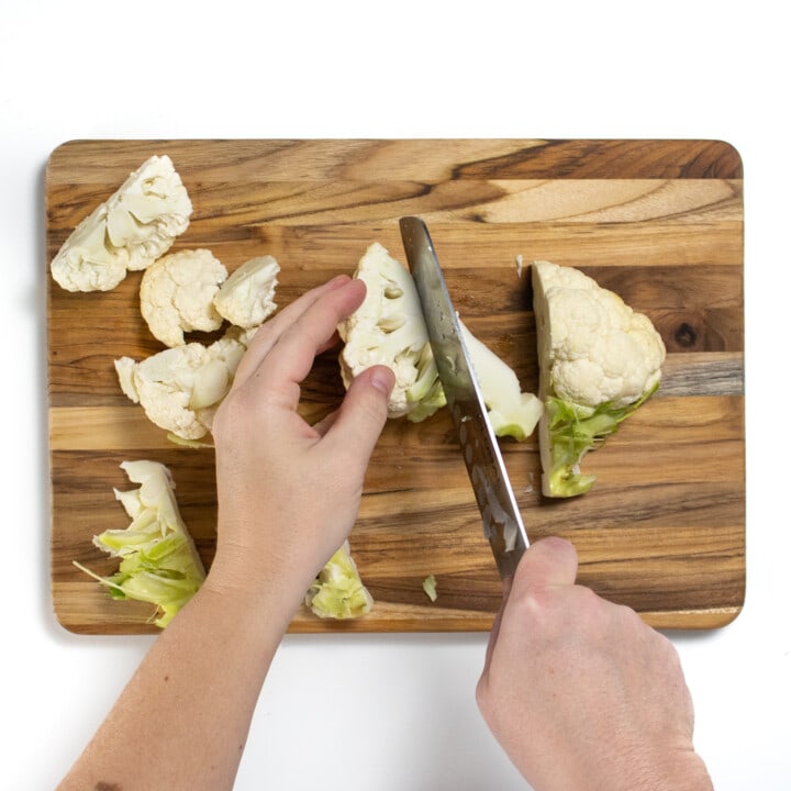 A wooden cutting board to hands are cutting florets of cauliflower.