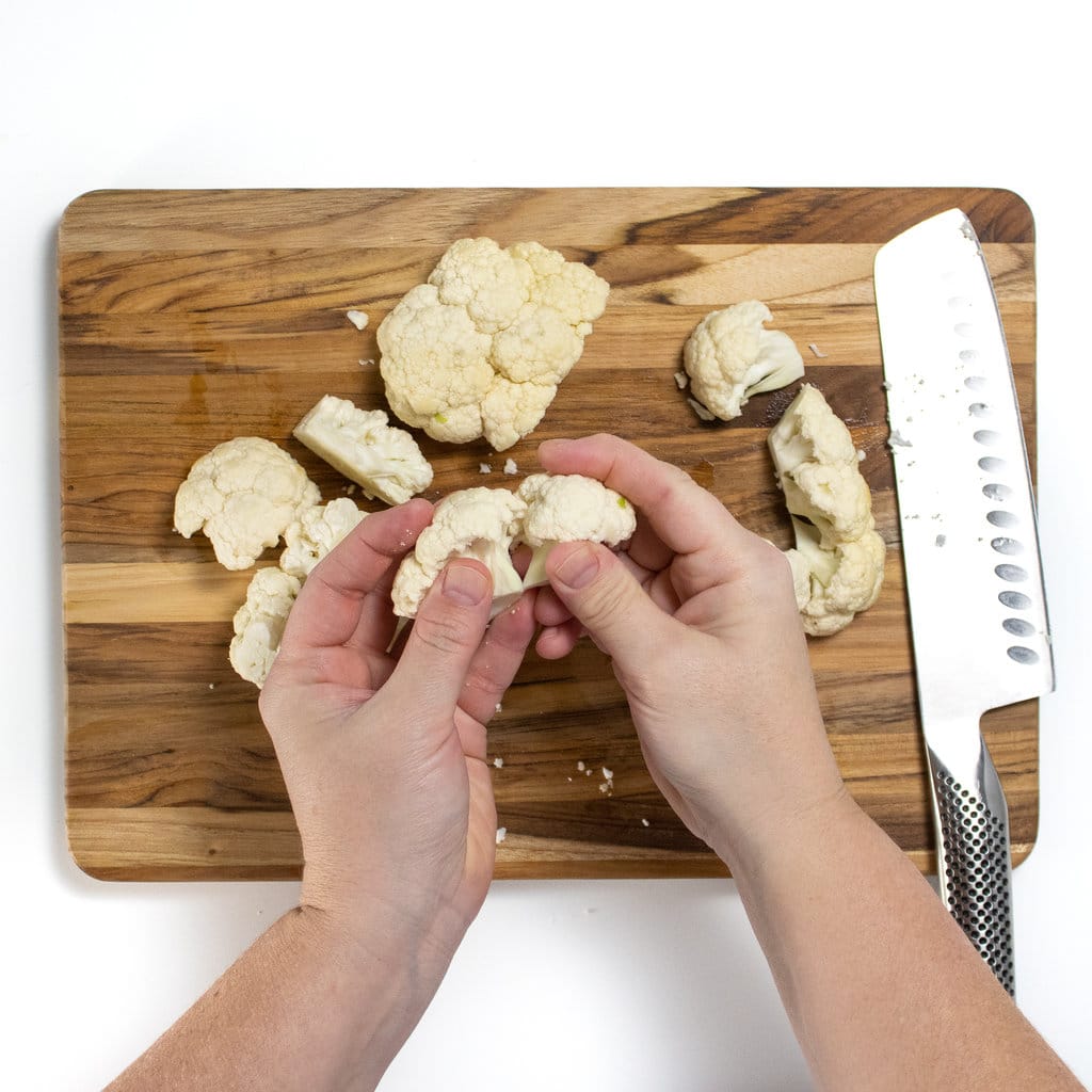 On a wooden cutting board against a white background two hands are breaking cauliflower head into florets.