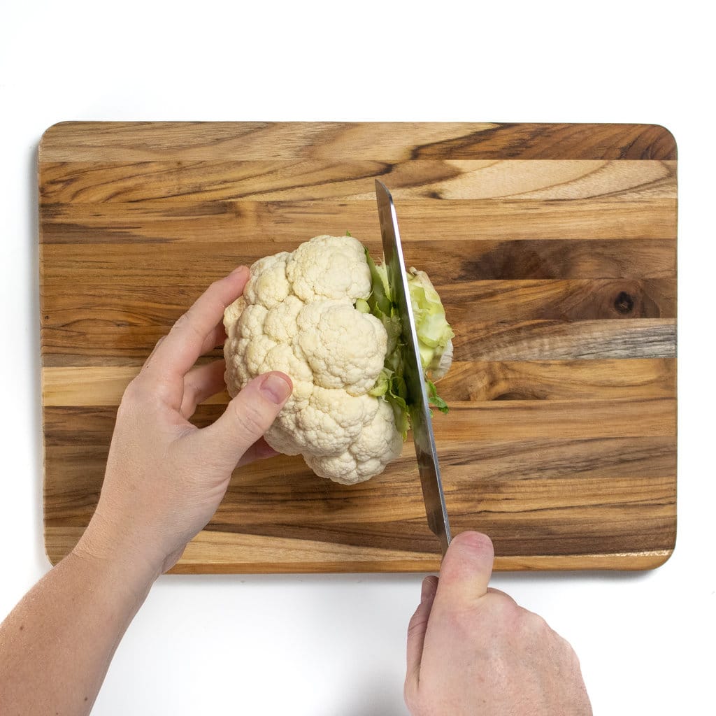 On a wooden cutting board and hand is holding a cauliflower head and the other hand is cutting off the base or stem of the cauliflower.