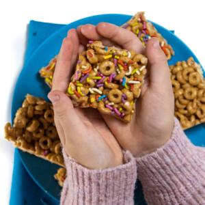 Two kids hands holding a cereal bar with colorful rainbow sprinkles against a plate of cereal bars.