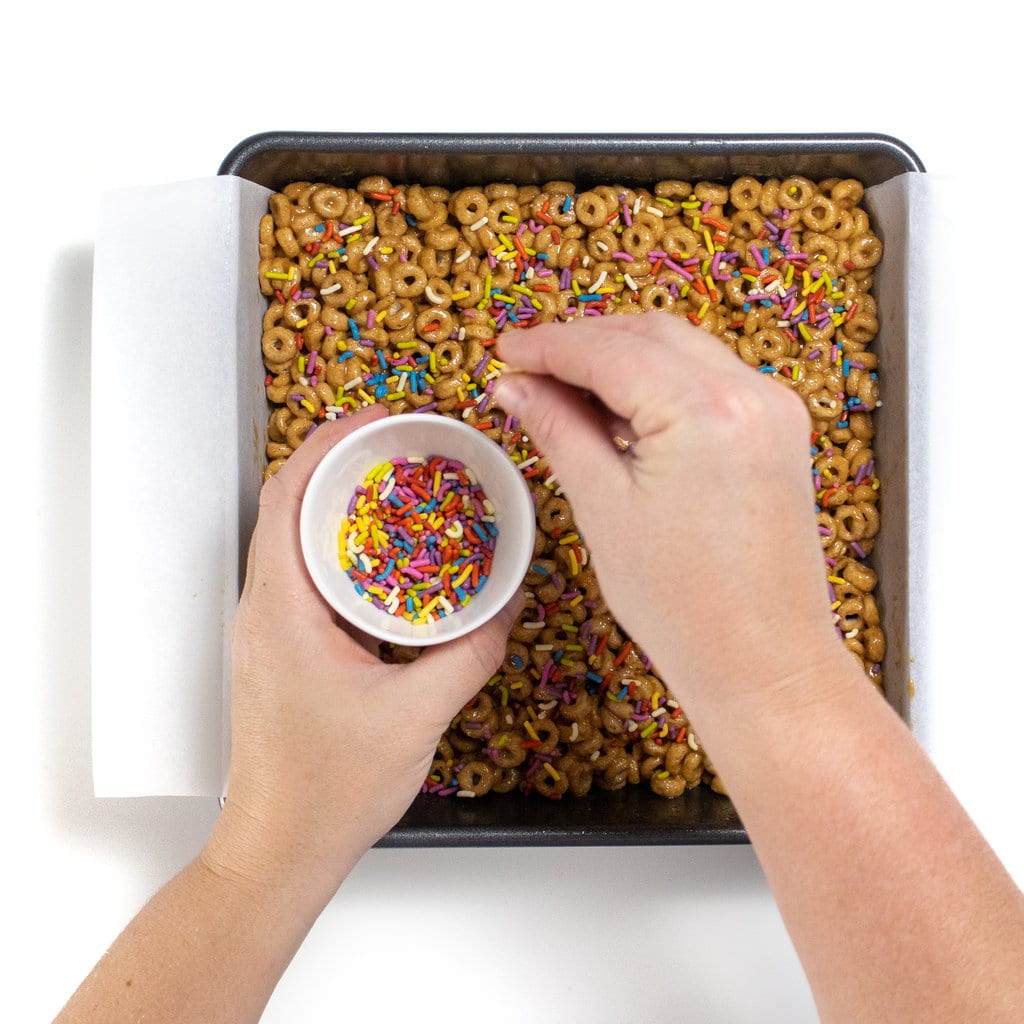 A square baking dish with cereal bars and hand sprinkling sprinkles over it.