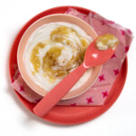 A pink kids bowl and plate with a pink napkin full of banana yogurt with a spoon resting on top.