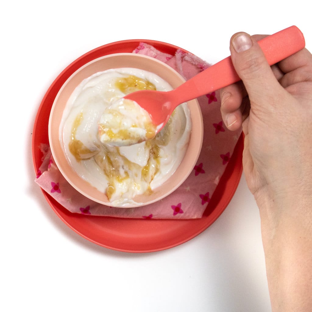 A hand holding a pink spoon over a pink kids bowl full of banana yogurt.