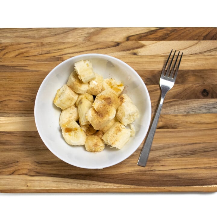 A cutting board with bananas, cinnamon and maple syrup with a fork sitting next to it.