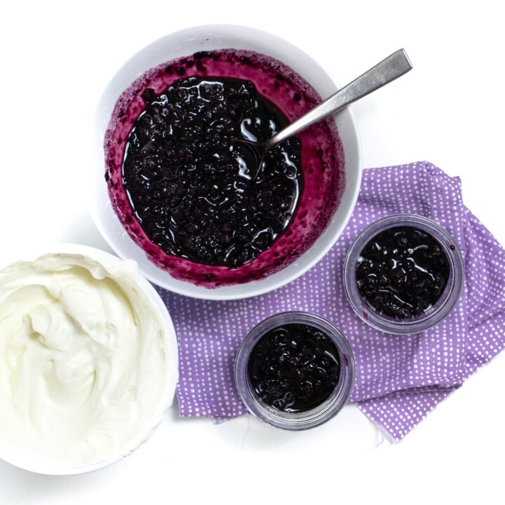 A bowl of blueberry compote and yogurt being put into small jars on a purple napkin against a white background.