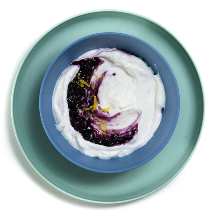 Blue kids bowl with a green kids played against a white background with blueberry yogurt with a sprinkle of lemon zest.