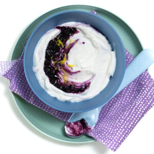 A white background with a teal kids plate with a blue kids bowl full of blueberry yogurt.