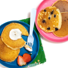 A blue and pink kids plate with yogurt pancakes on top some with blueberries on them and a side of strawberries.