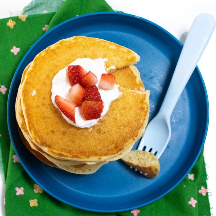 Blue kids plate and a green napkin with a stack of yogurt pancakes with a dollop of yogurt and chunks of strawberry on top.