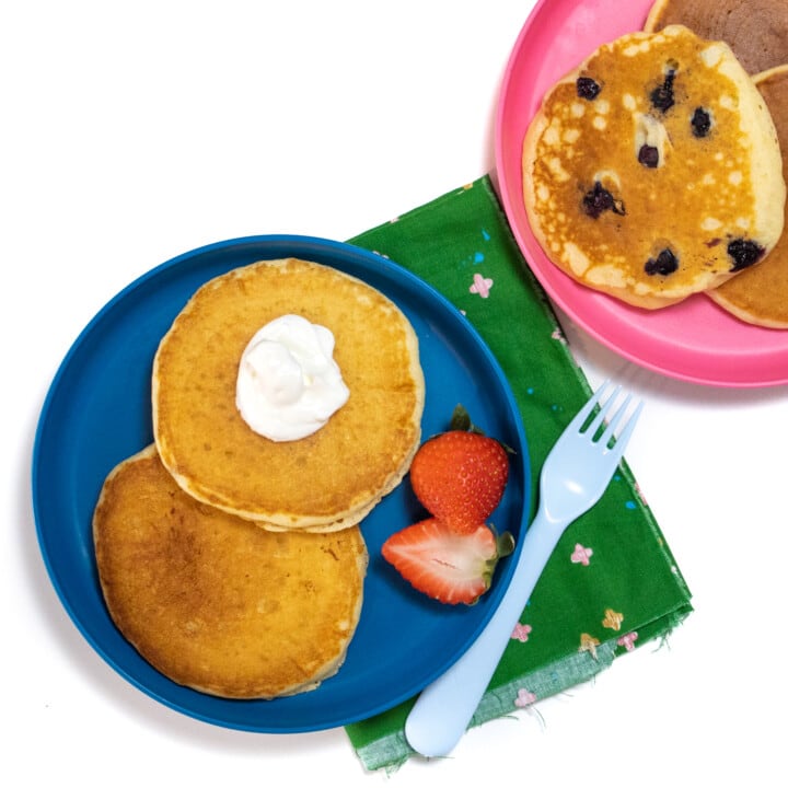 A blue and pink kids plate with yogurt pancakes on top some with blueberries on them and a side of strawberries.