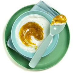 A blue kids bowl on top of a green kids plate with a mango yogurt inside with a spoon resting on top with mango yogurt in it.