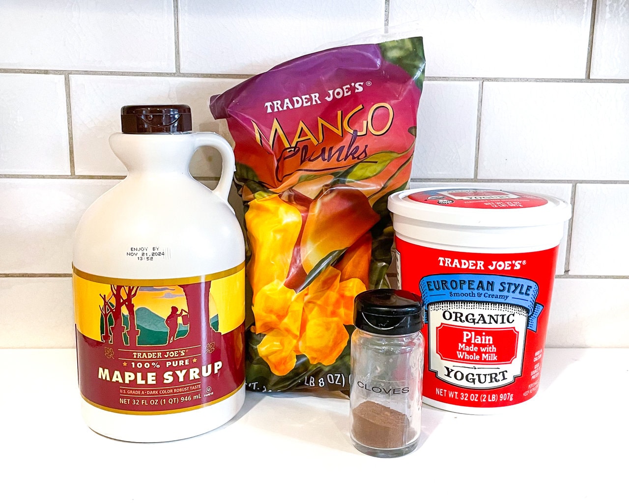 A white kitchen counter with the ingredients for the recipe – yogurt, frozen mango, nutmeg, and maple syrup.