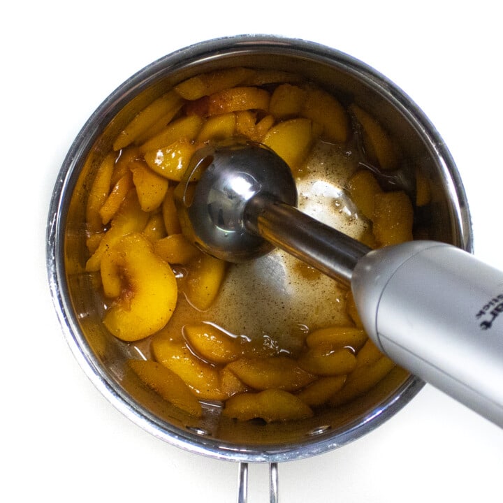 Silver saucepan with simmered peaches getting blended with an immersion blender.