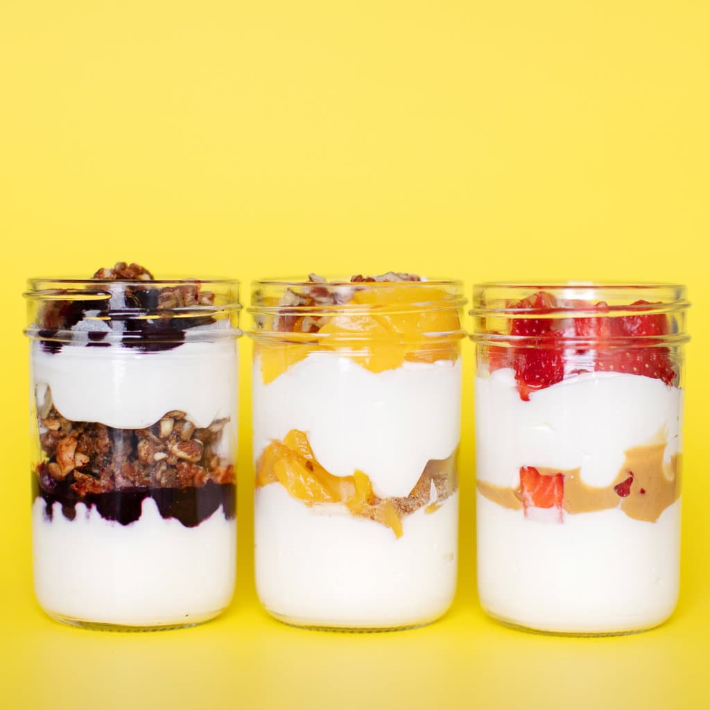 Three glass jars full of fruit filled yogurt parfait's for kids with lots of fruit and healthy toppings against a yellow background.