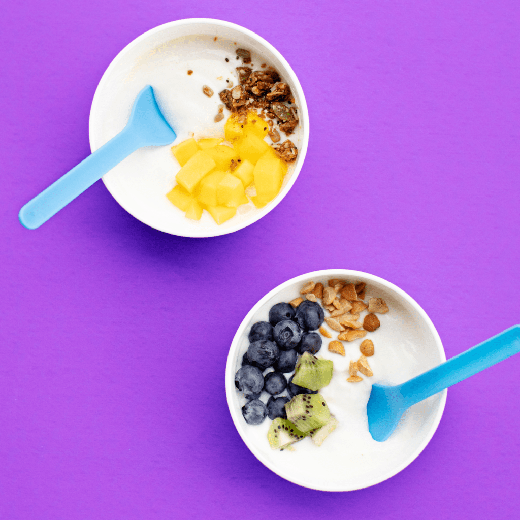Two white bowls full of yogurt and toppings against a purple background.