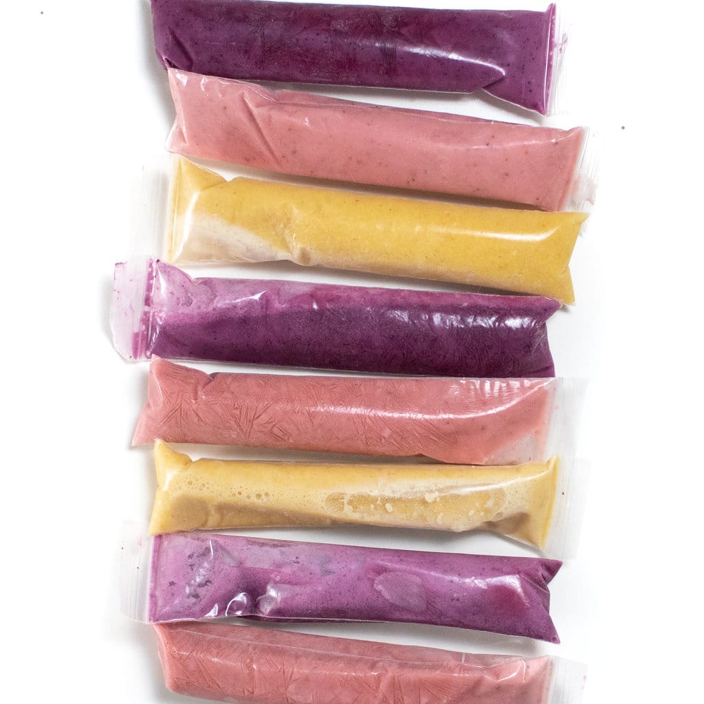 Tubes of yogurt and fruit frozen in blueberry, strawberry and peach are lined up in a row against a white back job.