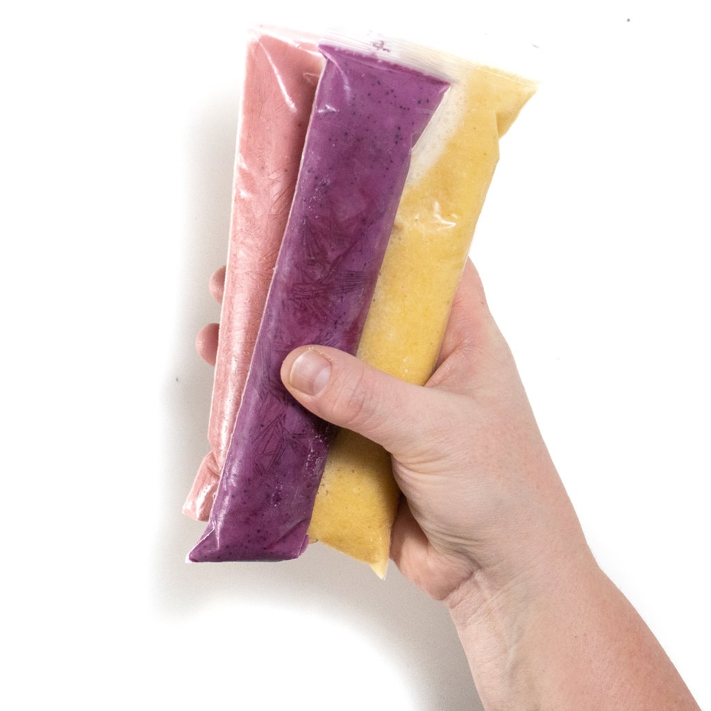A hand holding 3 different yogurt tubes in different flavors against a white background.