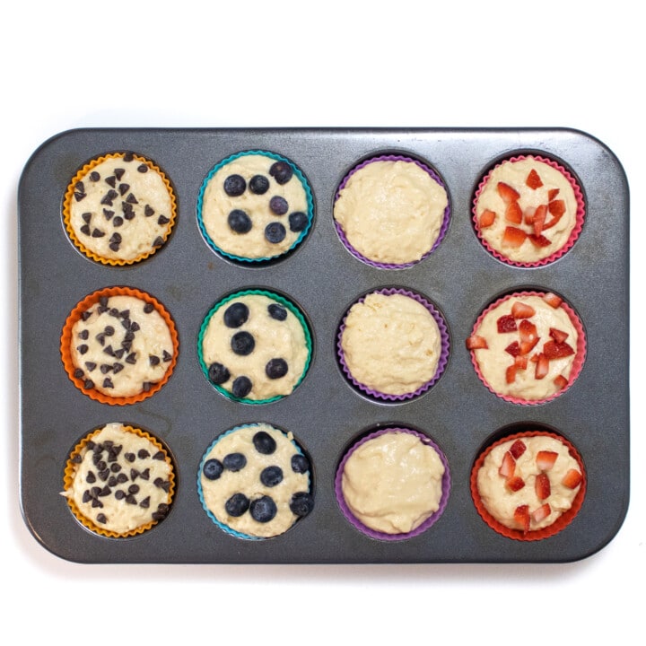 A muffin tin full of colorful liners, muffin batter and toppings.