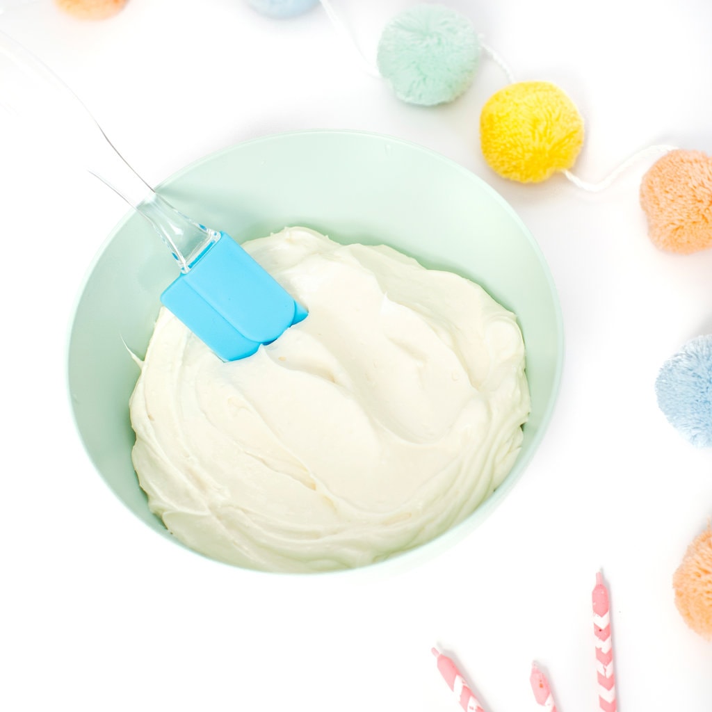 Teal green kids bowl filled with a smooth and healthy frosting against a white background with some colorful party props. 
