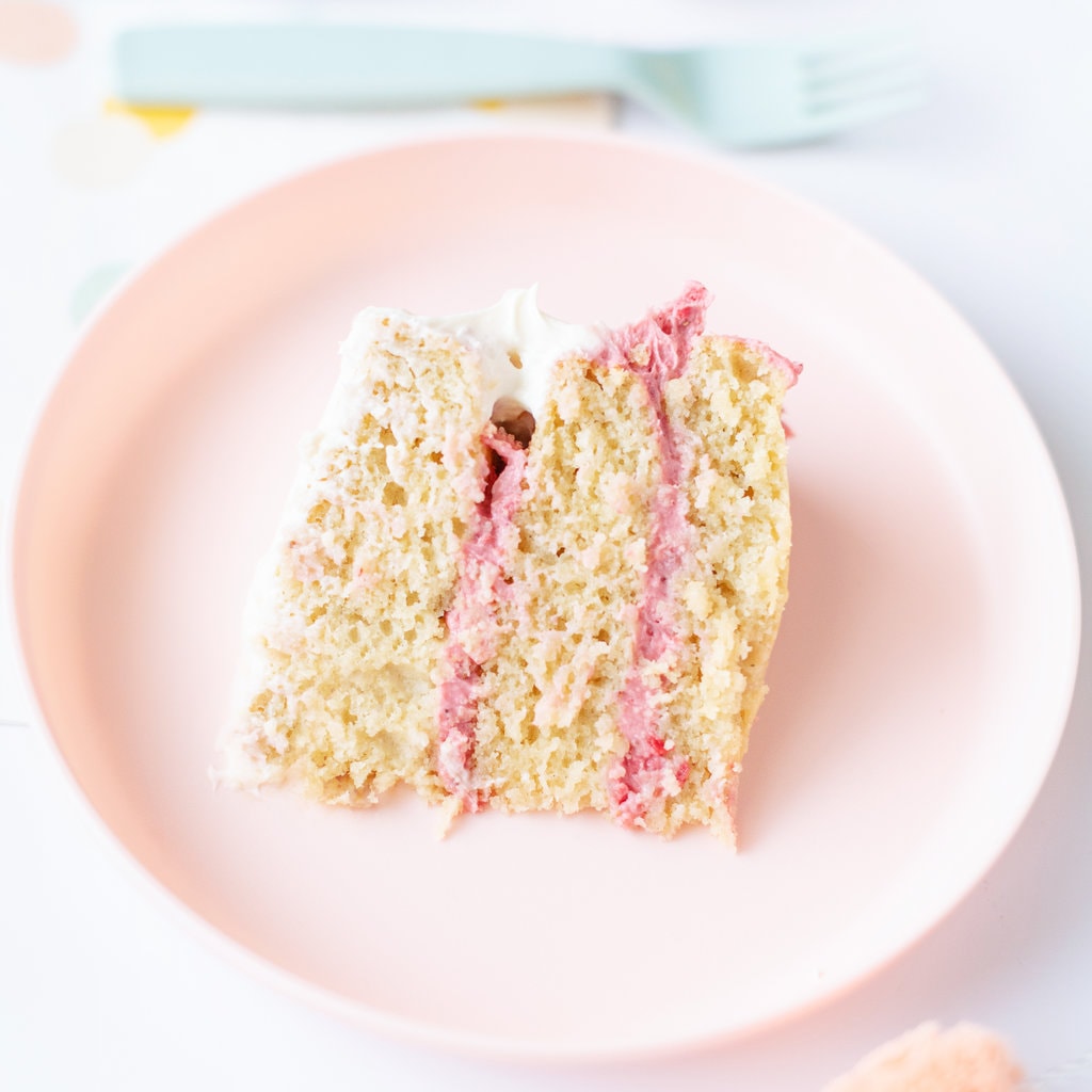 A piece of smash cake for baby with pink frosting between the layers on a pink kids plate with a blue fork.