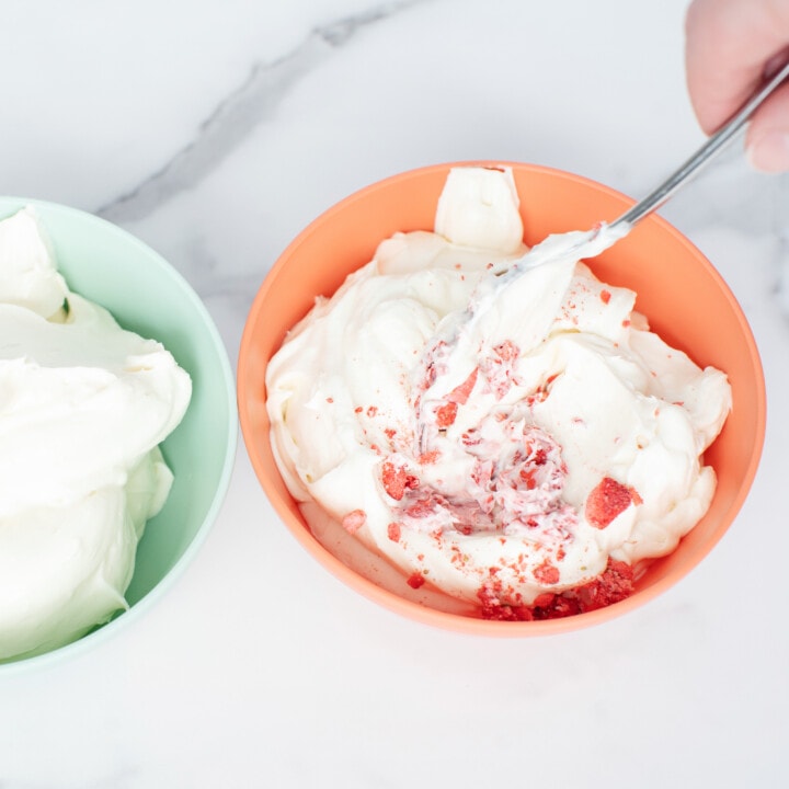 A hand mixing healthy cream cheese icing with dehydrated strawberry pieces in a pink bowl against a white background.