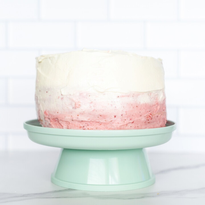 Up close of a ombre smash cake with white and pink frosting on a teal cake stand against a white background.