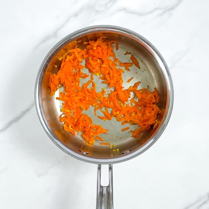 Silver saucepan on a marble surface with cooked carrots and garlic inside.