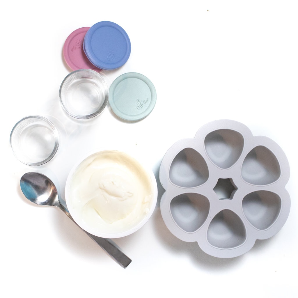 Spread of yogurt, ice cube tray, glass jars and lids against away background.