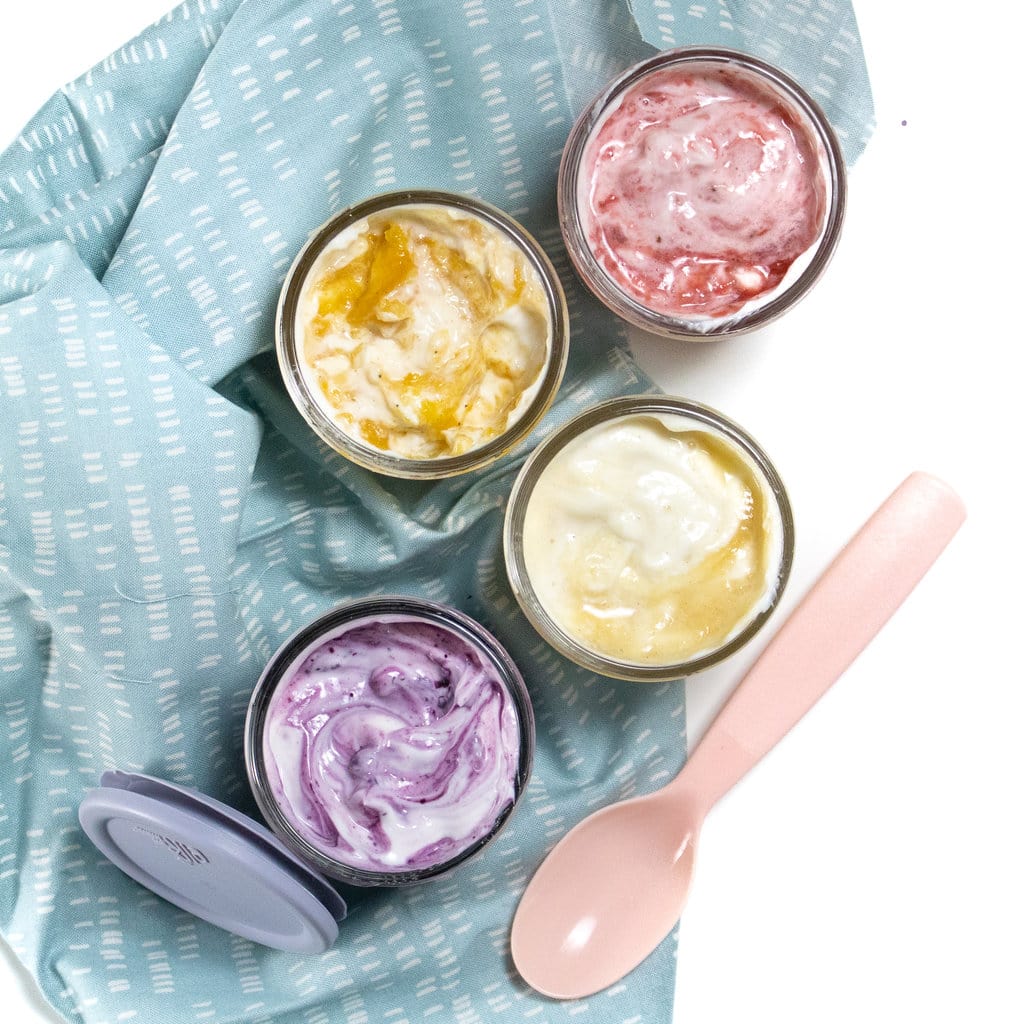 Four glass jars full of yogurt with fruit swirled on top with a blue napkin a pink spoon and a blue lid against a white background.