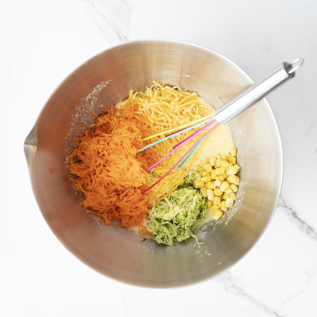 A silver mixing bowl and a marble countertop with grated carrots zucchini corn and cheese at four veggie muffins.