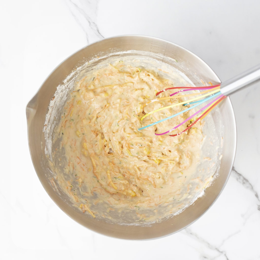 Silver mixing bowl with veggie muffin batter and a colorful whisk.