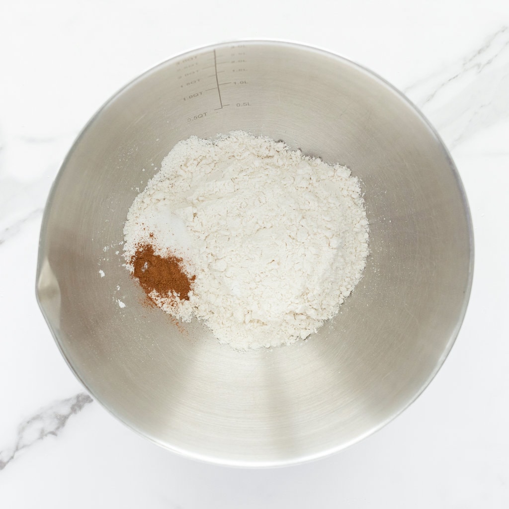 Silver mixing bowl with flour, baking powder, salt and cinnamon against a marble counter.