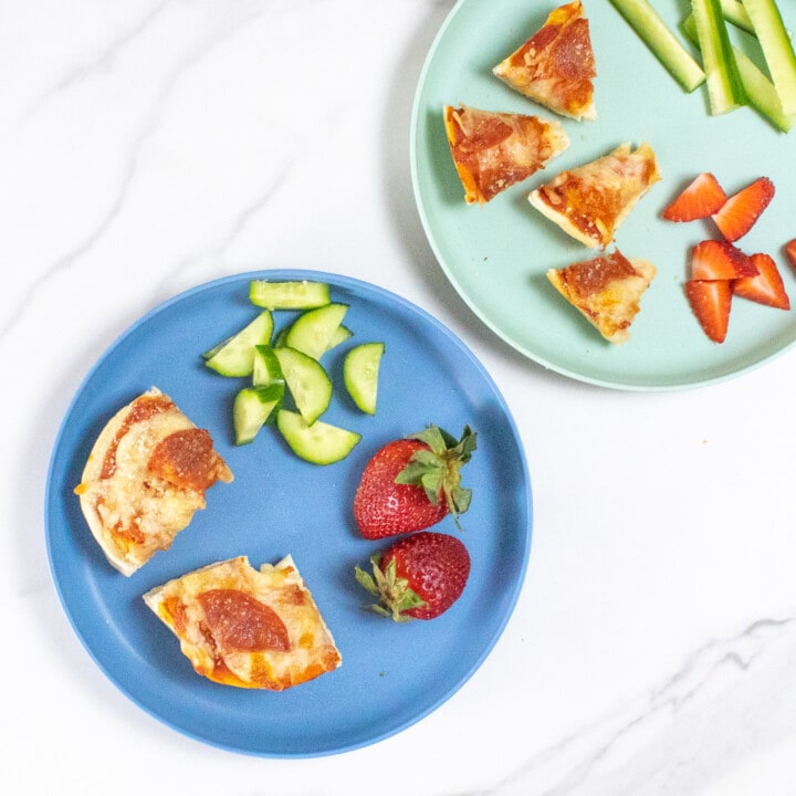 Two blue kids plates full of bagel pizza is cut into different sizes with cucumbers and strawberries on a marble countertop.