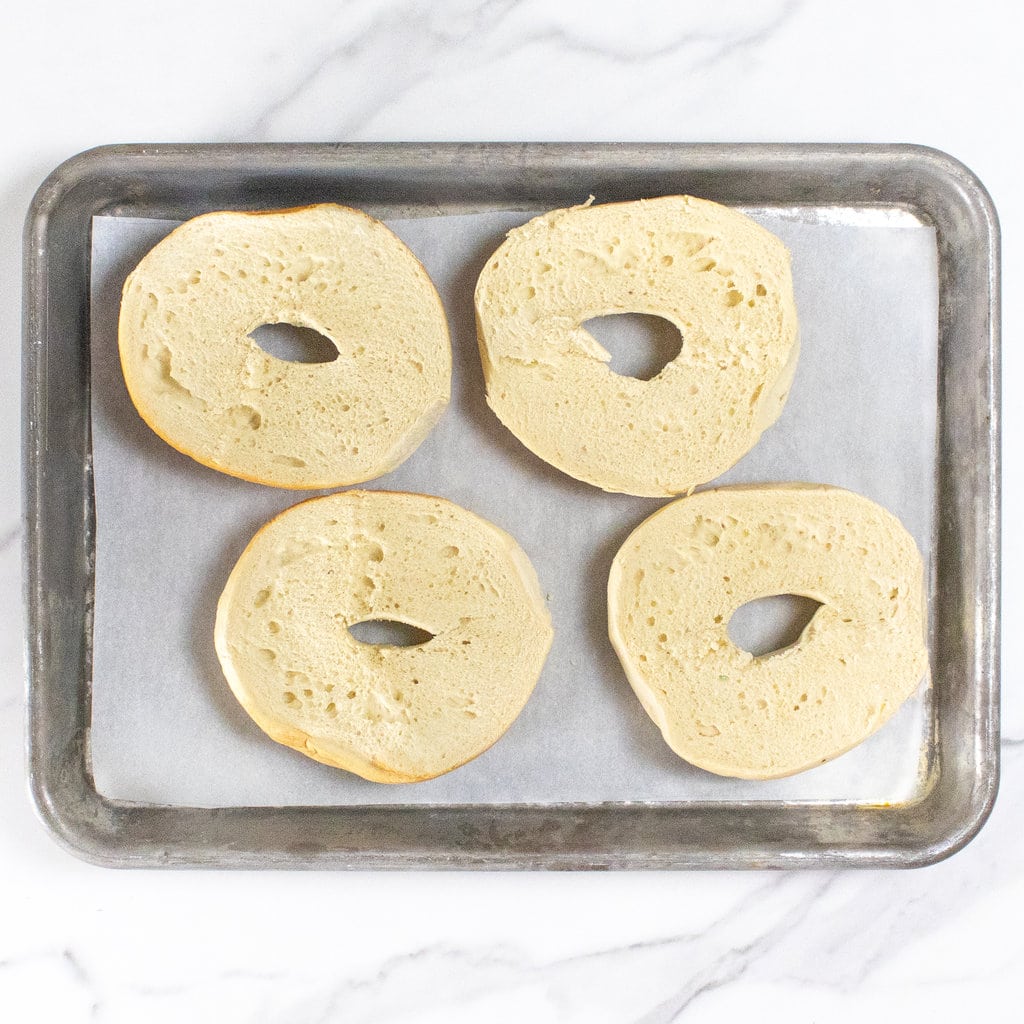 Bagels cut open on a baking sheet against a marble backdrop.