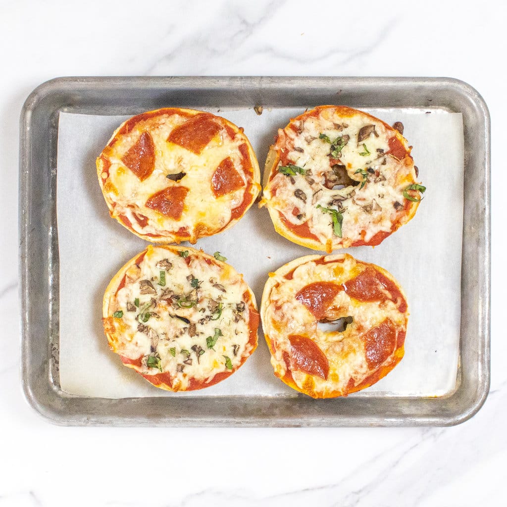 Four bagel pizzas on a baking sheet on a marble countertop with different toppings.