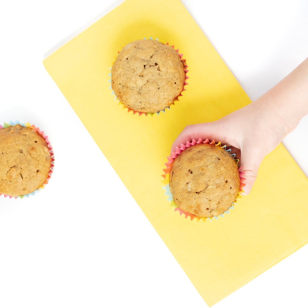 Kids hand reaching onto a yellow napkin with banana muffins in colorful wrappers.