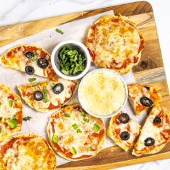 A cutting board with different toppings of English muffin pizzas with a small bowl of Parmesan and basil to sprinkle on top.