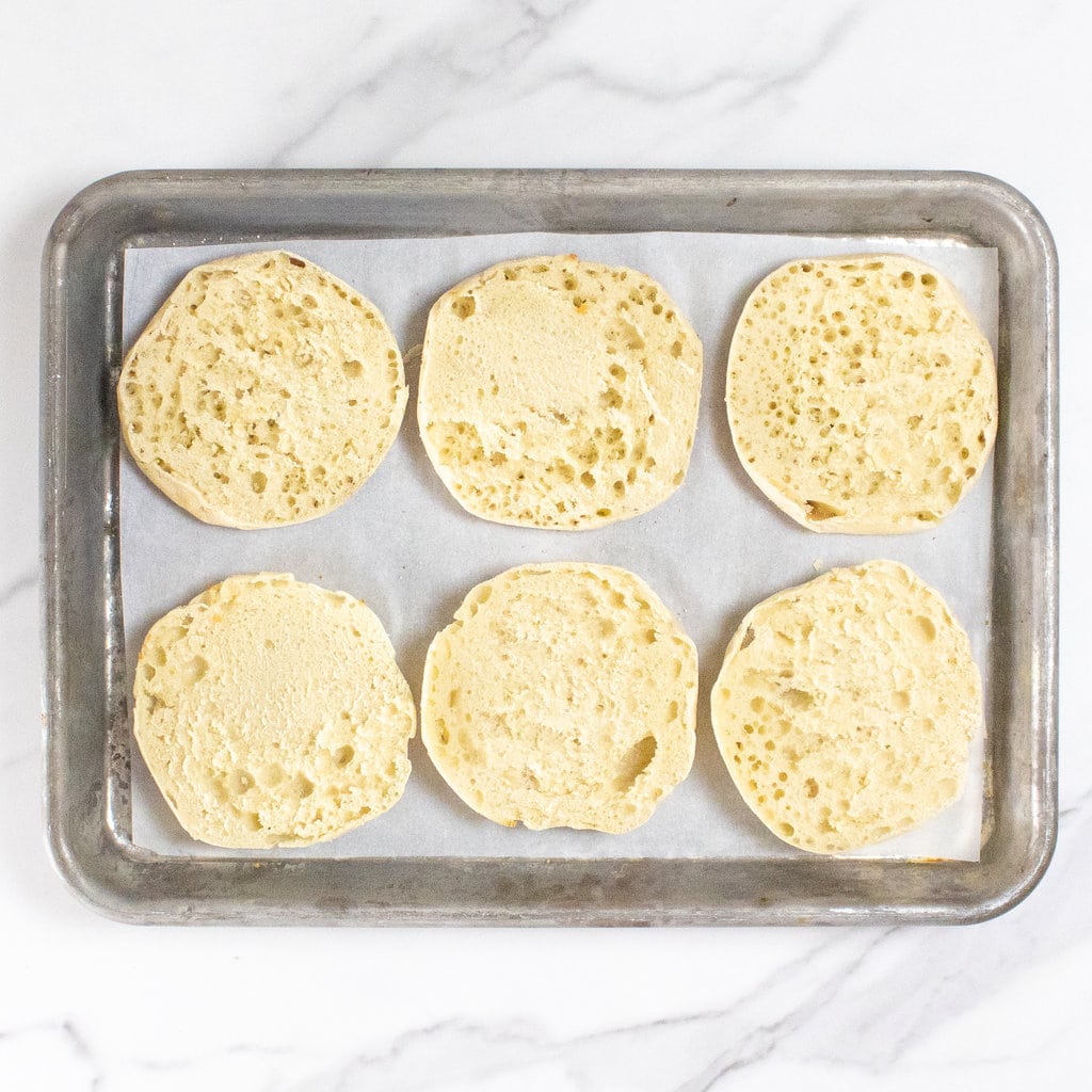 a baking sheet on a marble countertop with sliced English muffins on top of parchment paper.
