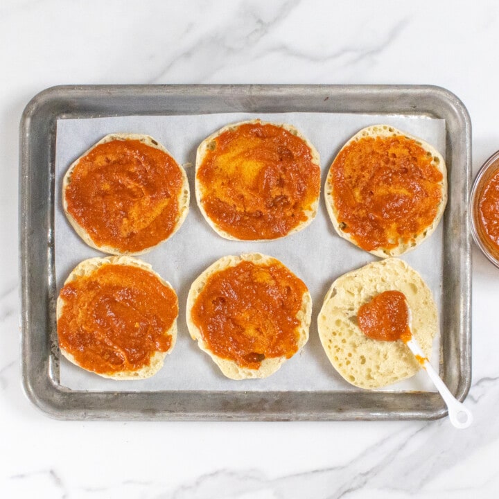A baking sheet on a marble countertop with six English muffins with pizza sauce on top.