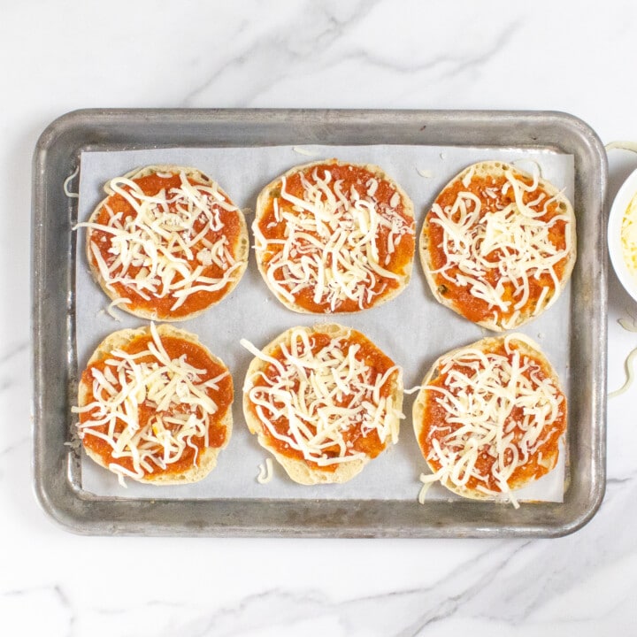 A baking sheet on a marble counter top with English muffins with sauce and cheese sprinkled on top.