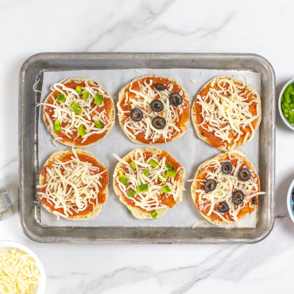A baking sheet and a marble countertop at six English muffins with various toppings.