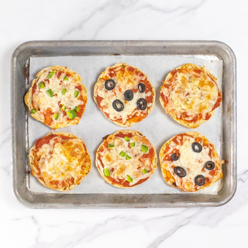 A baking sheet on a marble countertop with six cooked English muffin pizzas with different toppings.