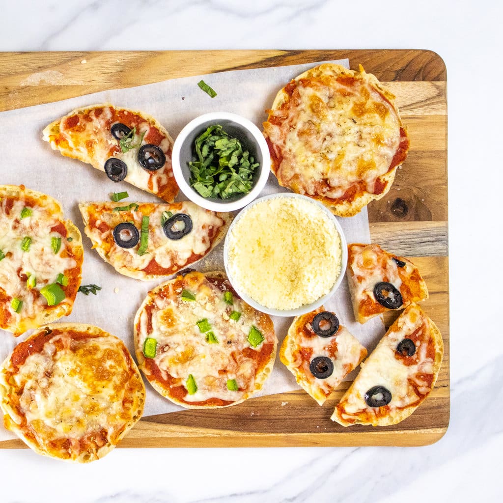 A wooden cutting board on a marble countertop with several different English muffin pizzas with different toppings, a bowl full of Parmesan and spicy basil for topping.