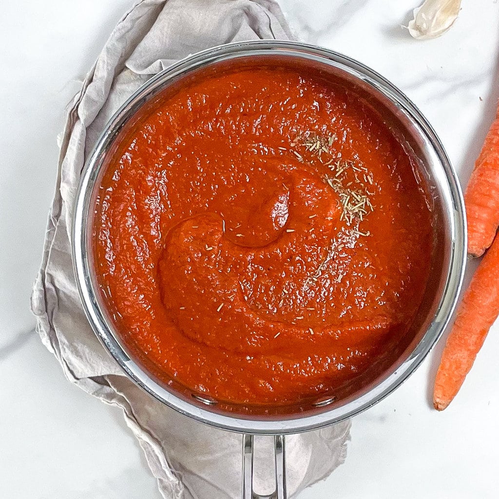 Silver saucepan with red pizza sauce and a tan dishcloth and carrots and garlic scattered around.