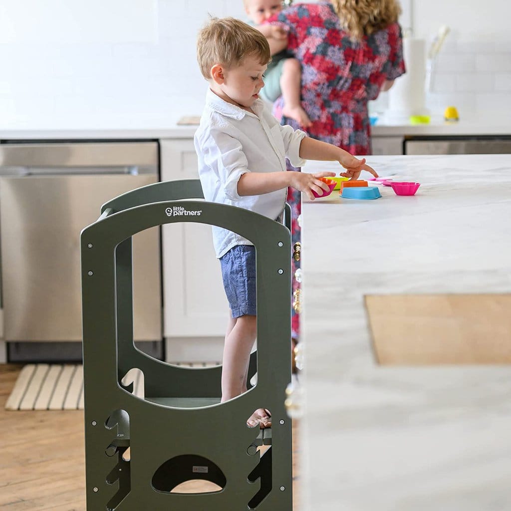 White and bright kitchen with a boy standing on a learning tower playing with some toys on the counter. 