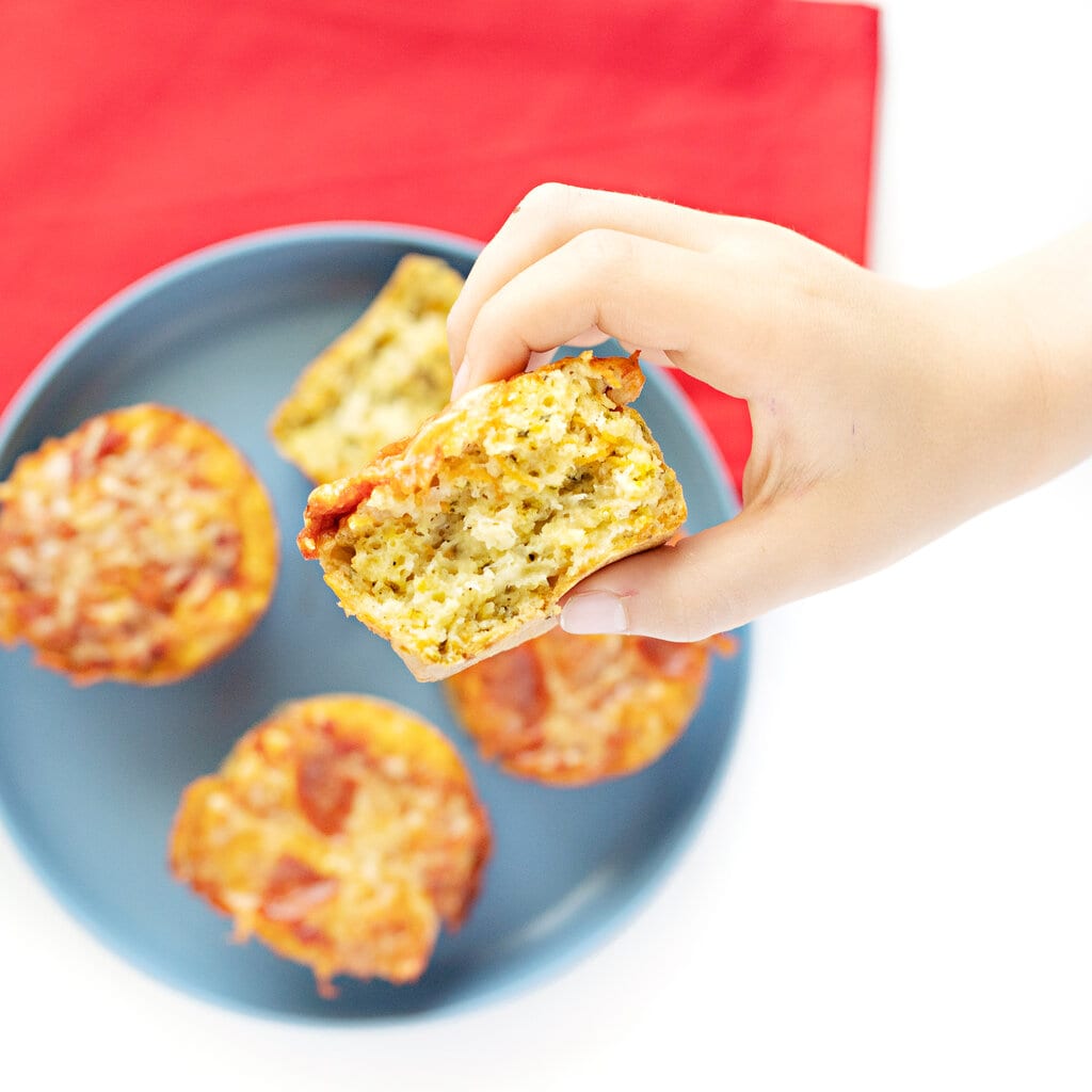 A hand holding a pizza muffin over a plate of pizza muffins with a red napkin against a white background.